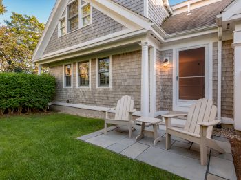 A house with gray shingle siding and a small, neatly maintained lawn. Two Adirondack chairs and a small table sit on a stone patio near a side door.