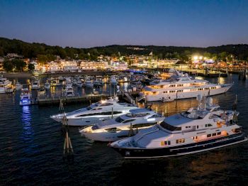 A serene night view of a marina with several yachts docked, lights reflecting on the water, and a vibrant waterfront community in the background.