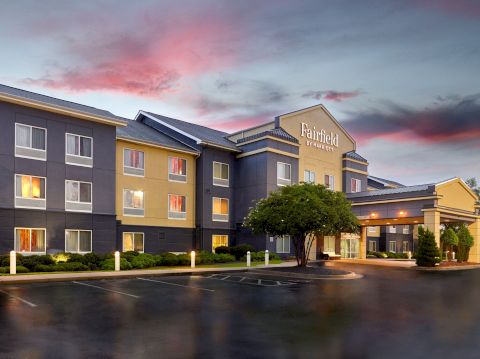 A modern three-story hotel building with "Fairfield" signage, exterior lighting, and landscaped surroundings, under a pink and blue sky at dusk.