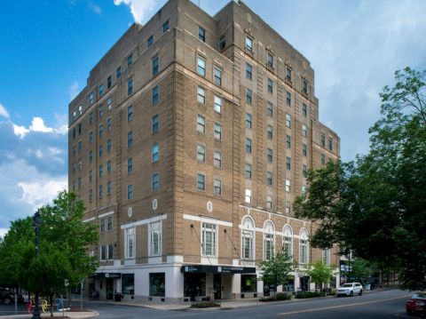 A large, multi-story brick building with street-level shops, surrounded by trees and cars, under a partly cloudy sky.