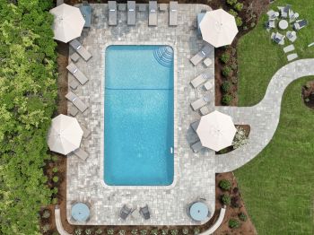 An overhead view of a rectangular pool surrounded by lounge chairs, umbrellas, and greenery, with a curved path leading to a separate seating area.