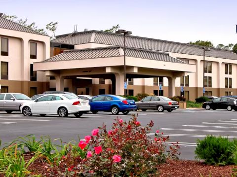 An image of a hotel with a parking lot in front, featuring several cars and landscaped area with flowers and shrubs.