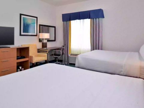 The image shows a hotel room with two beds, a TV on a stand, a desk and chair, a lamp, and a window with curtains.