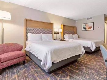 The image shows a hotel room with two double beds, a red armchair, a floor lamp, nightstand with lamps, and wall art. The decor is modern and comfortable.