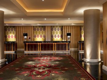 The image shows an elegant hotel lobby with a richly patterned carpet, large pillars, subdued lighting, and a reception desk in the background.