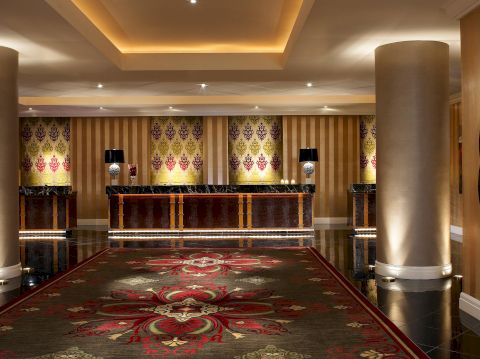 The image shows an elegant hotel lobby with a richly patterned carpet, large pillars, subdued lighting, and a reception desk in the background.