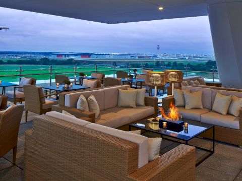 The image shows a modern outdoor lounge area with wicker furniture, cushions, and a firepit, overlooking a lush landscape with distant buildings.