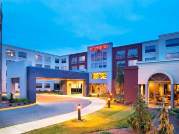 The image shows the exterior of a Sheraton hotel building, featuring a driveway entrance, illuminated signage, and a well-maintained landscape.