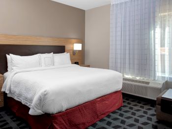 The image shows a neatly made bed with white linens in a hotel room, featuring a bedside table, lamp, wooden headboard, and a window with sheer curtains.