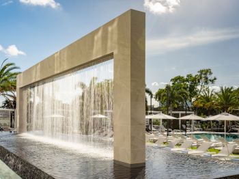 A pool area with loungers, umbrellas, and palm trees, featuring a large architectural waterfall structure under a sunny sky.