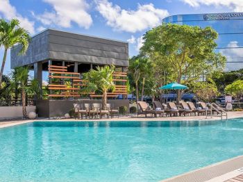 A pool area with lounge chairs, a shaded structure, and trees; a modern building is in the background under a blue sky with clouds.