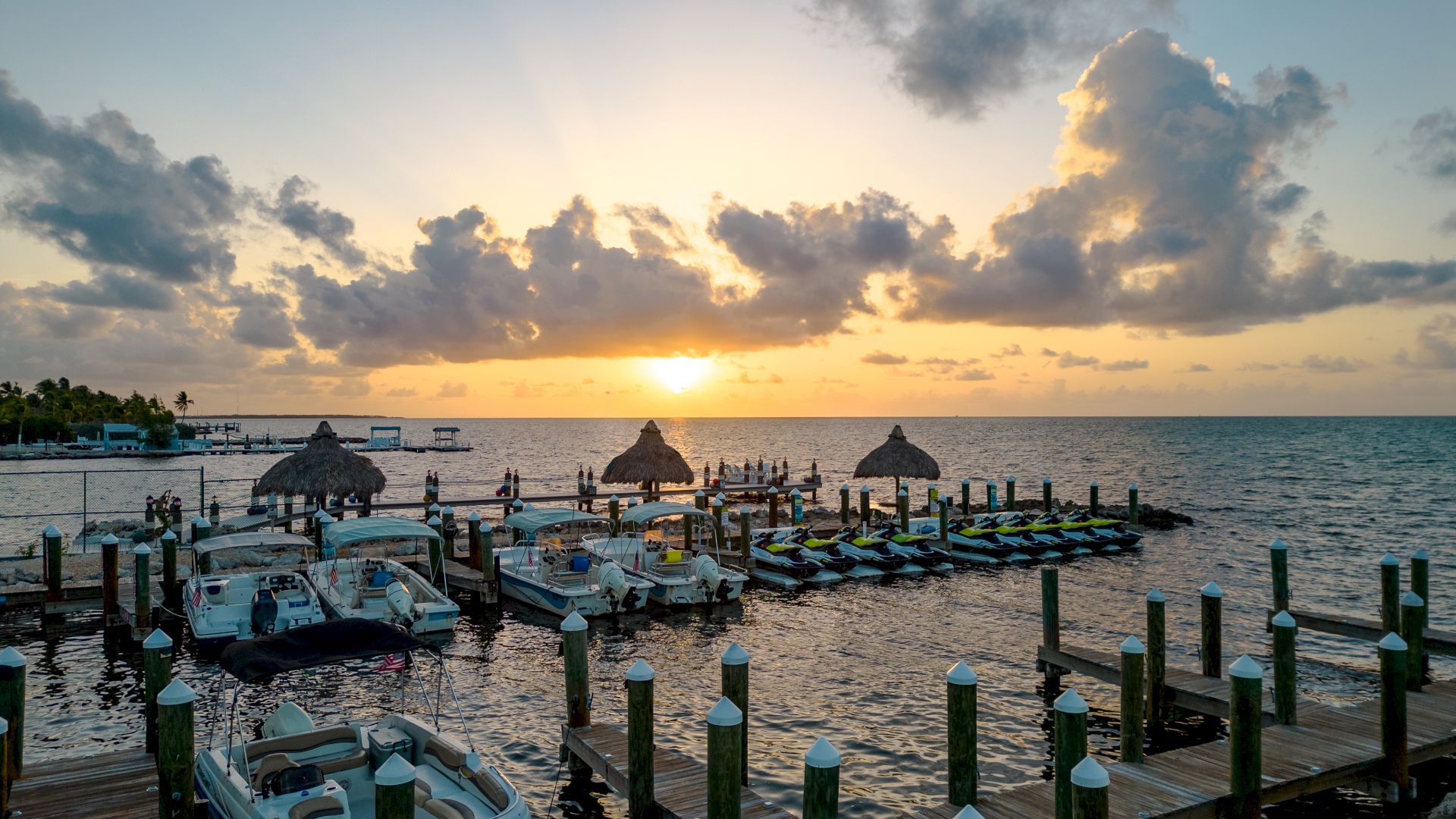 The image shows a serene marina at sunset with boats docked along piers and thatched-roof structures over the water.