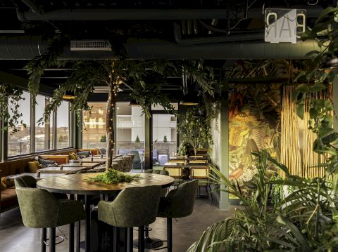 A cozy, modern restaurant with lush greenery, wooden decor, large windows, and comfortable seating arrangements, creating a serene dining atmosphere.