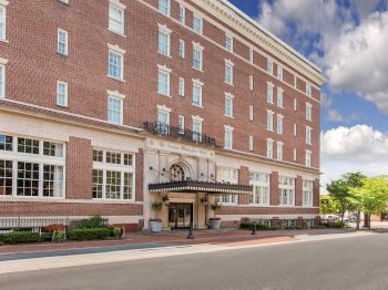 The image shows a multi-story brick building with large windows, an entrance canopy, and trees on the sidewalk on a clear day.